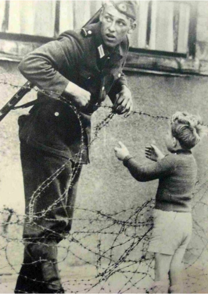 "Żołnierz NRD ignorujący rozkazy zabraniające przechodzenia przez mur berliński i pomagający młodemu chłopcu przedostać się na drugą stronę do jego rodziny. 1961 rok."